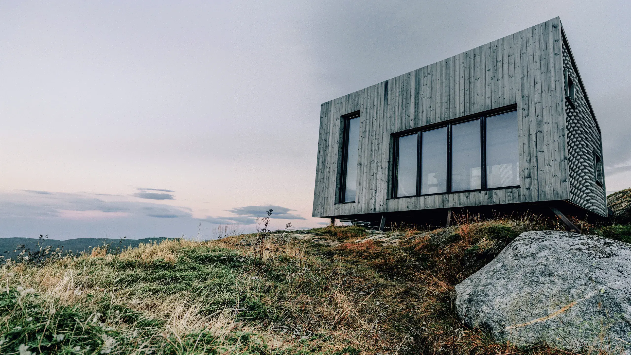 Moderne norsk hytte med grå trekledning og store svarte vinduspartier på eksponert fjelltopp – Natre vinduer